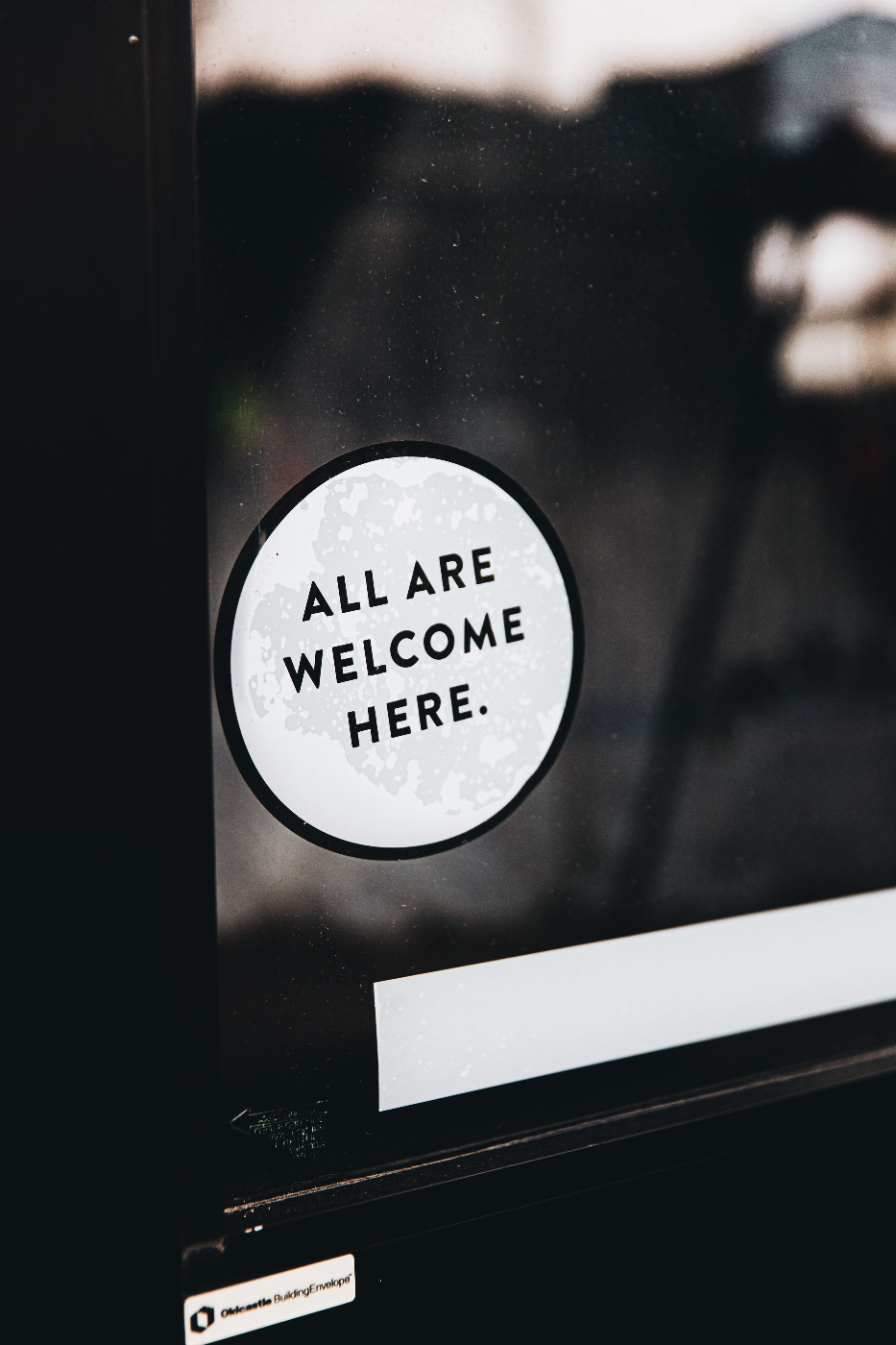 A shop door with a small round sign reading: All Are Welcome Here