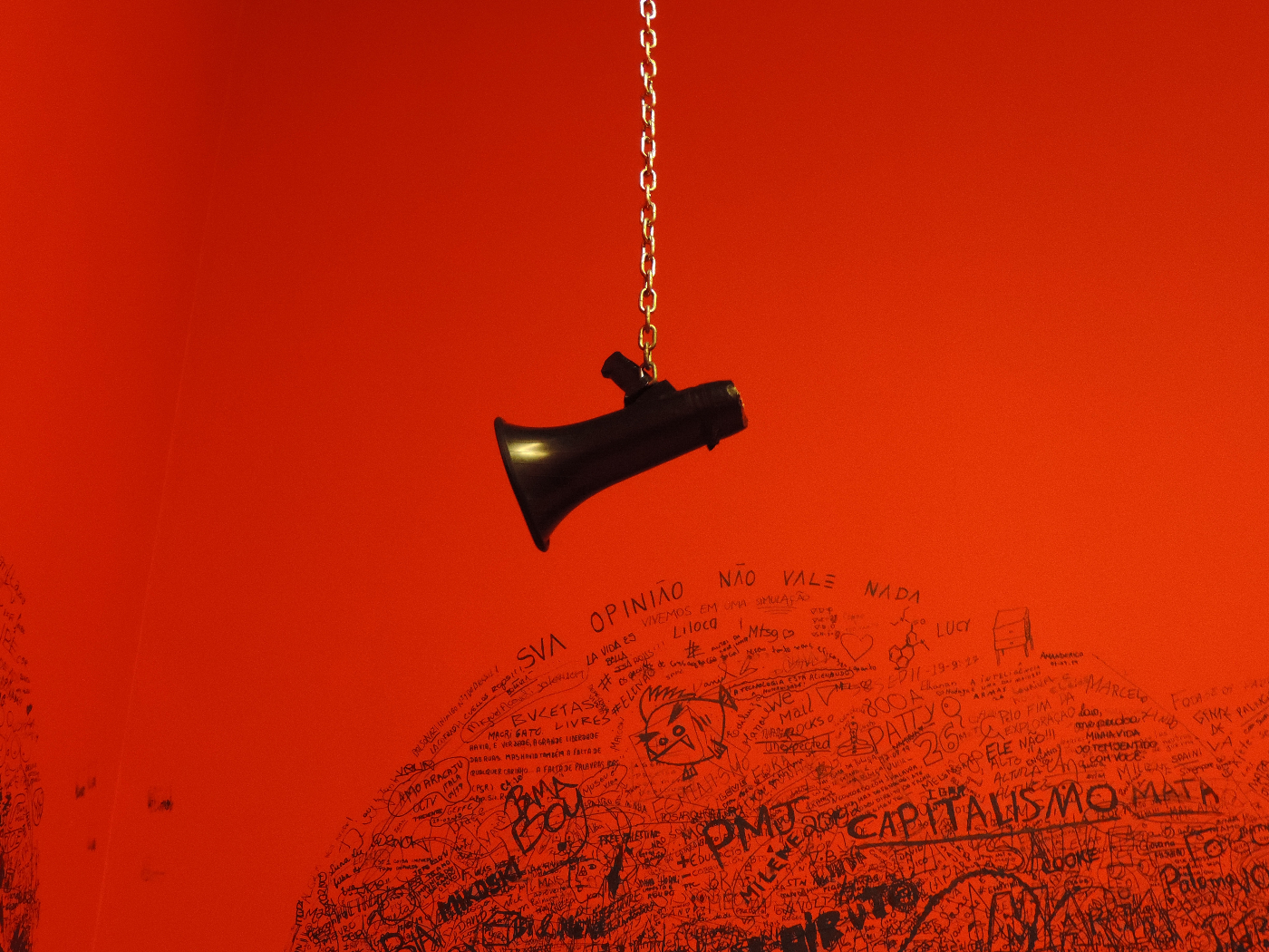 A magaphone hanging at the end of a chain against a red graffitied wall
