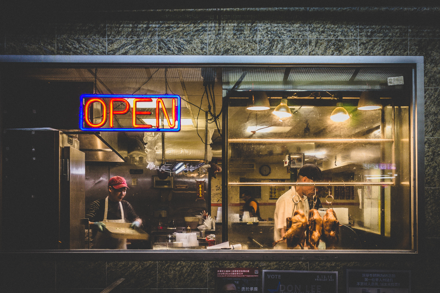 a small restaurant with roasted chickens in the window, an open sign and two men working inside