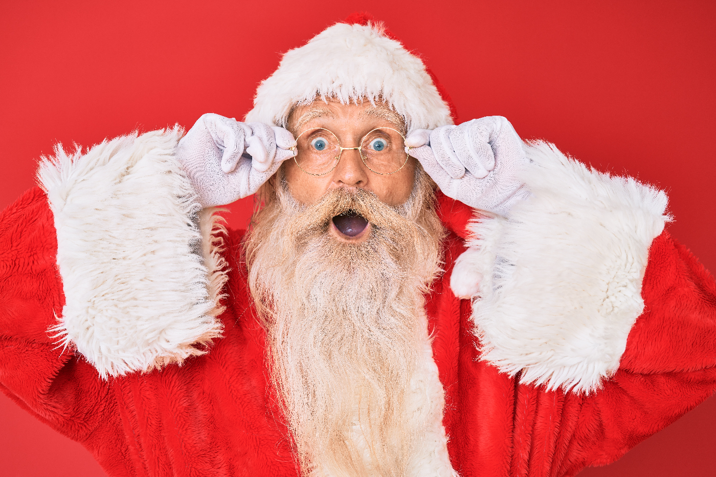 A man dressed as Santa with a natural beard, holding his round glasses and giving a open mouth smile