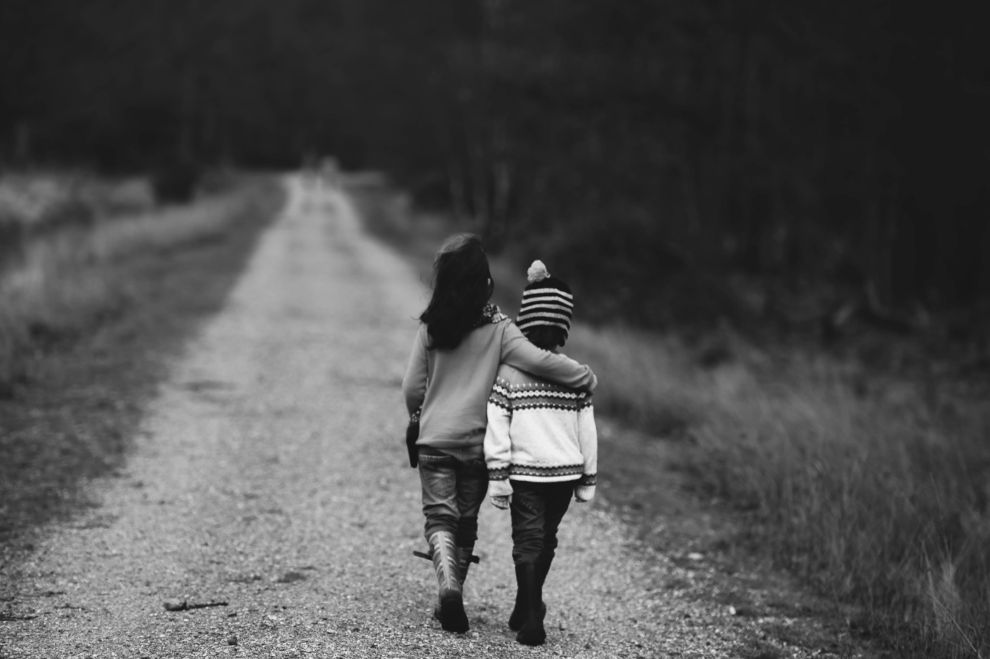 Two small children walking down a country road, one has her arm around the shorter one's shoulders