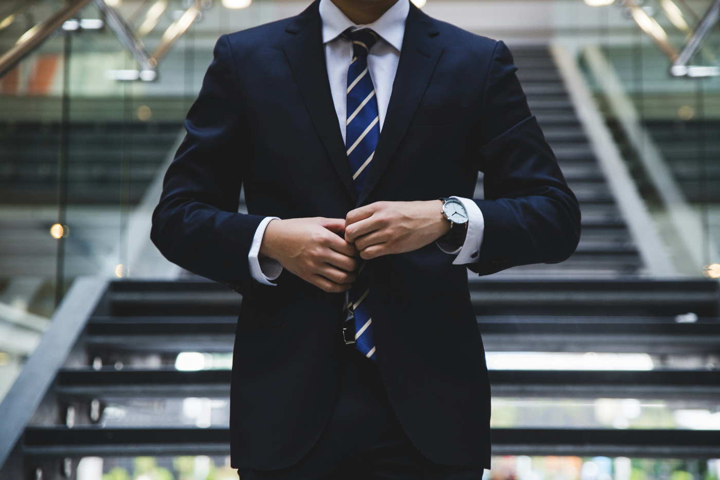 torso shot of a guy buttoning the jacket of his blue suit while standing in front of a long stairway