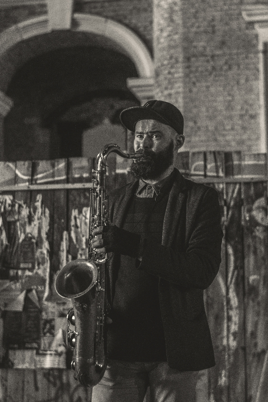 A bearded black man in ball cap, jacket and fingerless gloves playing sax in front of a graffitied wood fence