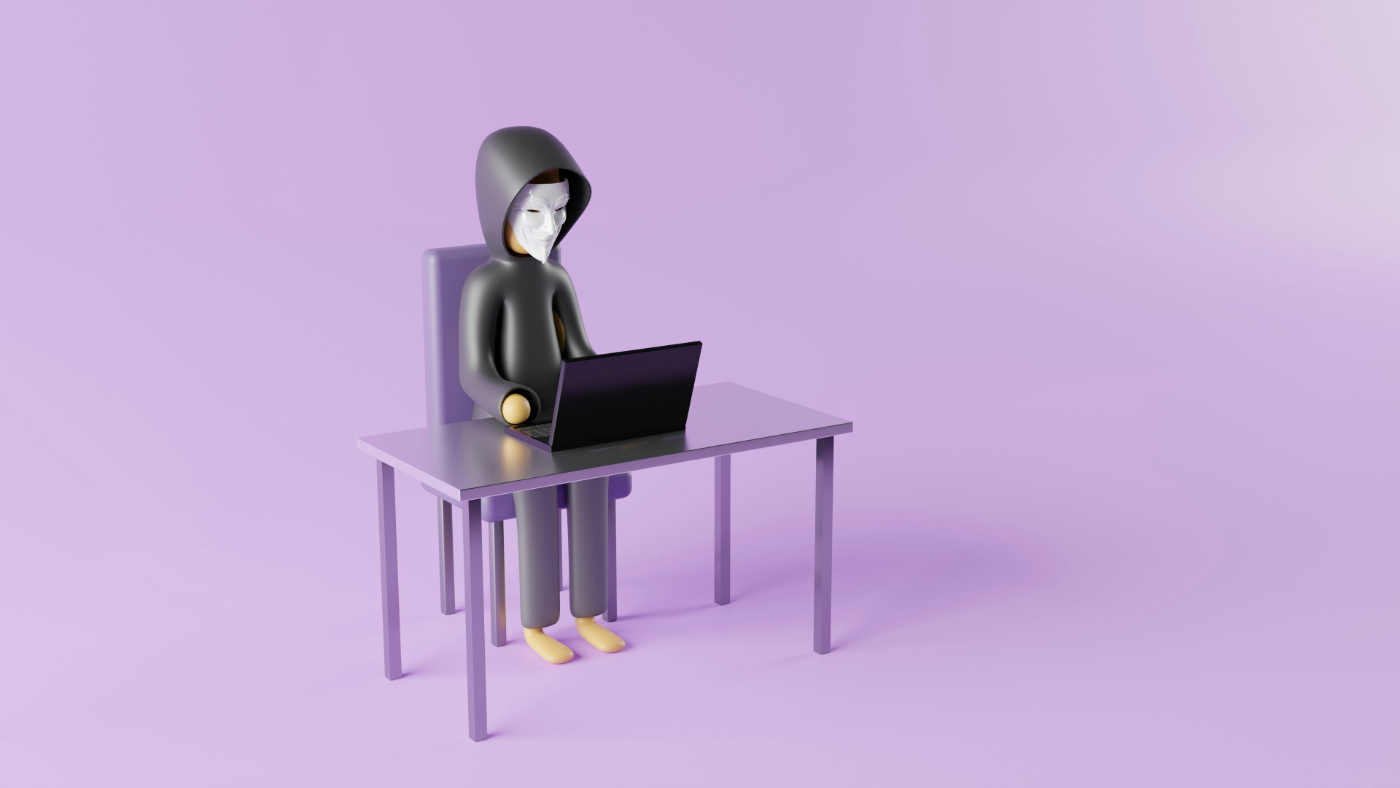 A toy man with an anonymous mask sitting at a small desk looking at a tiny computer