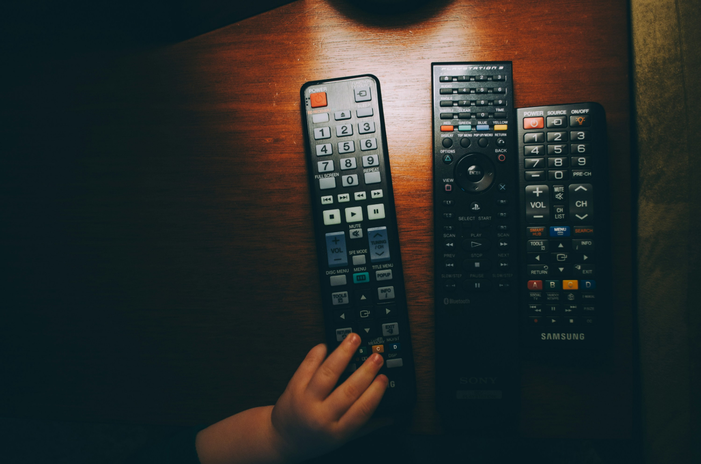 A child's hand touching one of three large media remotes