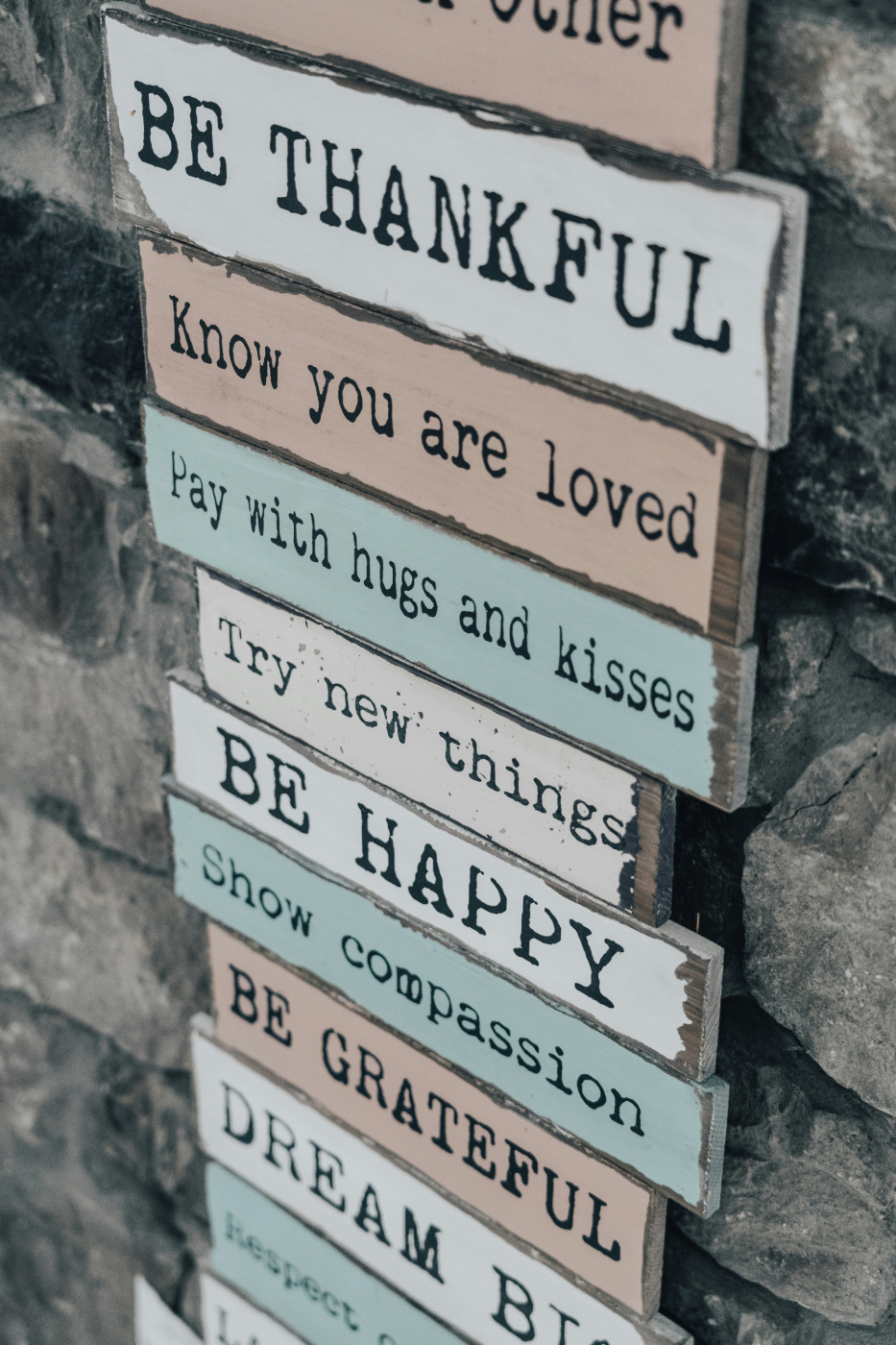 A sign board on a stone wall reading: Be Thankful, Know You Are Loved, Pay with Hugs and Kisses, and more