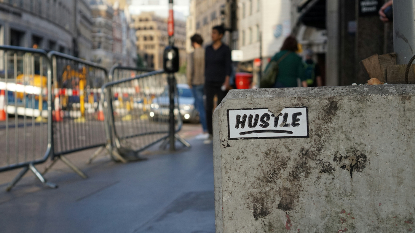 A city street and a torn sticker on a concrete barrier that says Hustle
