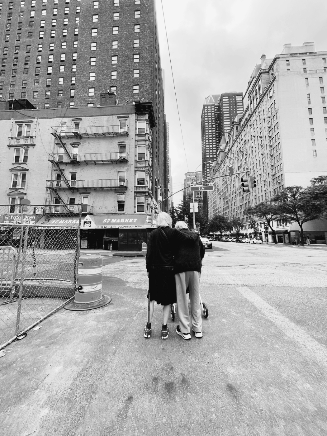 A gray scale image of an older couple supporting each other as they walk down a city street.