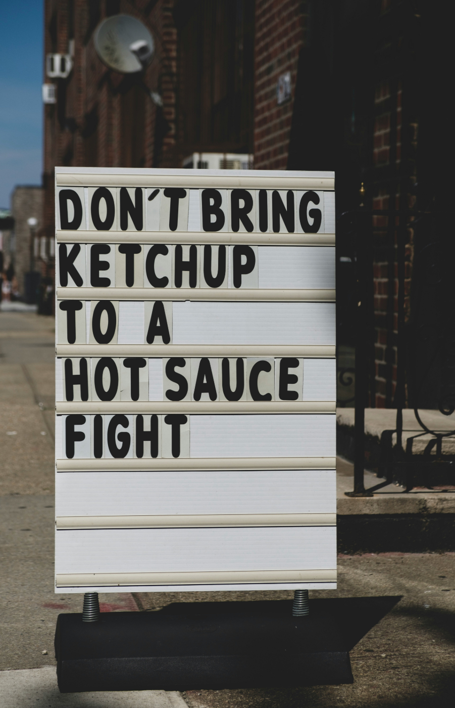 A sign on a sidewalk reading Don't Bring Ketchup to a Hot Sauce Fight