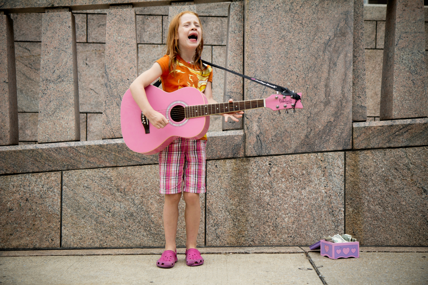 A red haired little girl with a pink guitar busking on the street