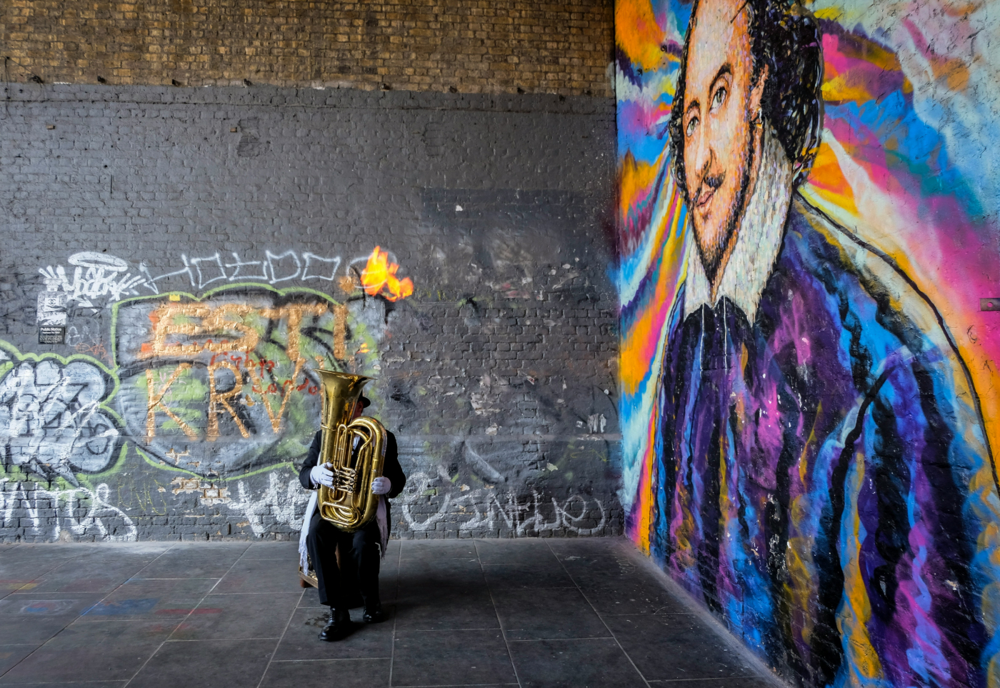 A man on the street in top hat and tails playing a tuba in front of a wall that has been graffitied with a portrait of Shakespeare in many colors