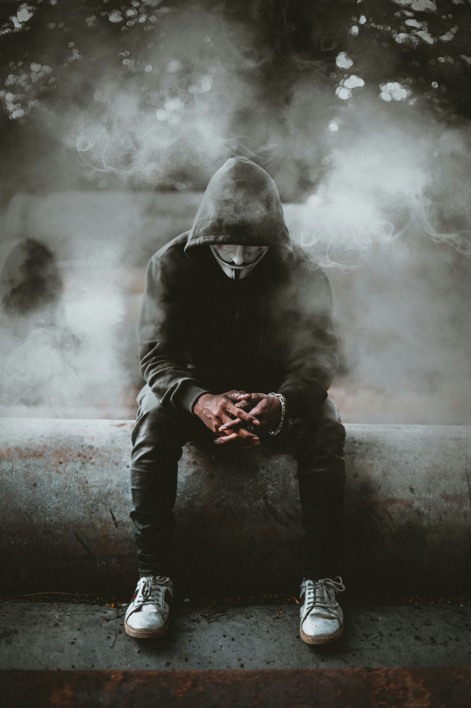 A man in a black hoodie and black pants wearing a V for Vendetta mask, sitting on the edge of a fountain