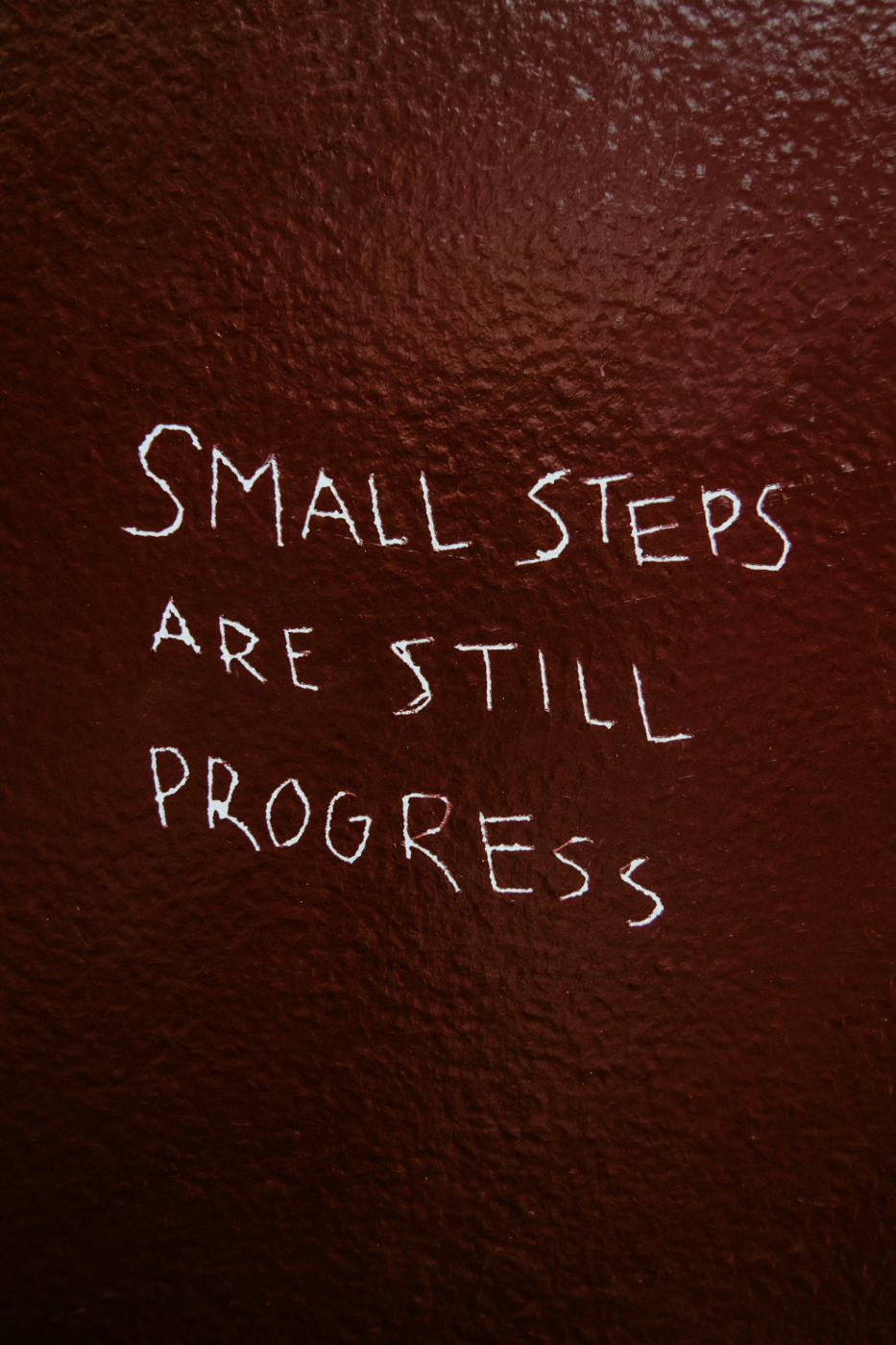 Letters scratched into a red painted wall reading, "Small Steps are still progress."