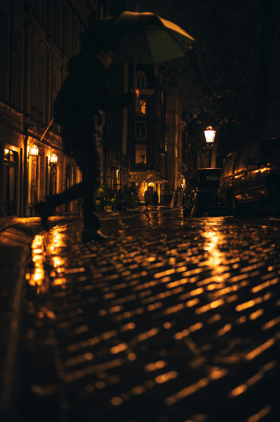 A barely visible man walks rainy cobblestone streets lit by street lamps