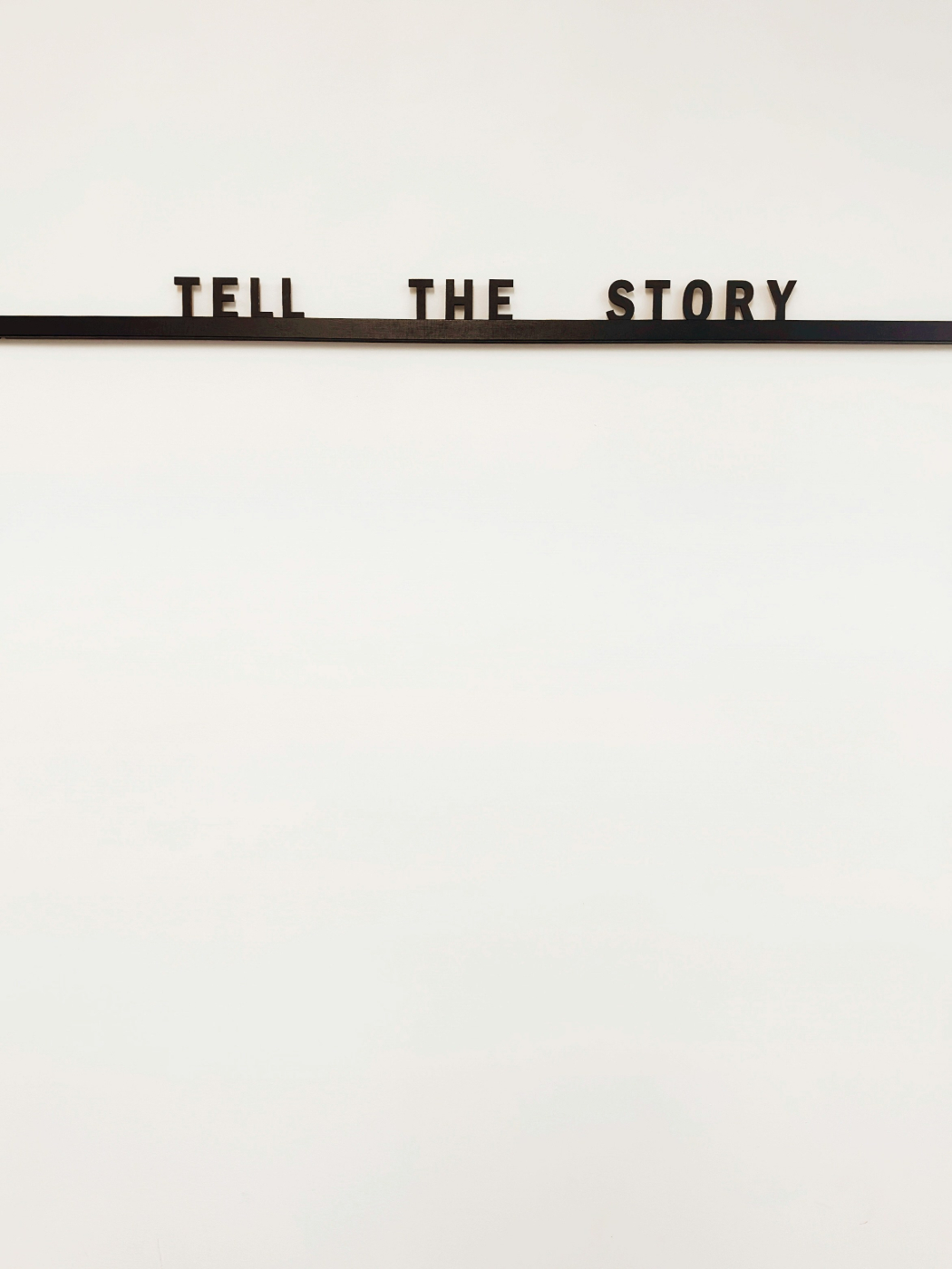 A blank wall with a thin self and the words Tell The Story on the shelf