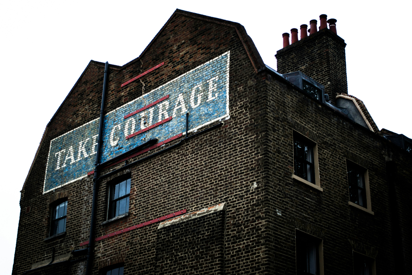 An old brick building with a blue and white sign painted on the side reading: Take Courage