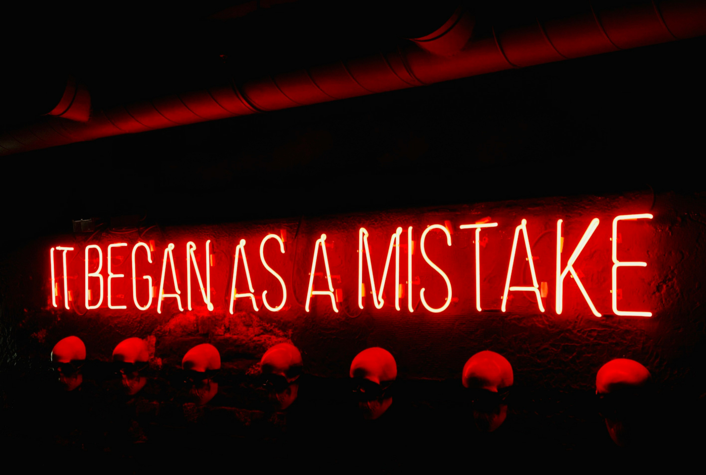  red neon sign over a row of skulls reading It Began As A Mistake