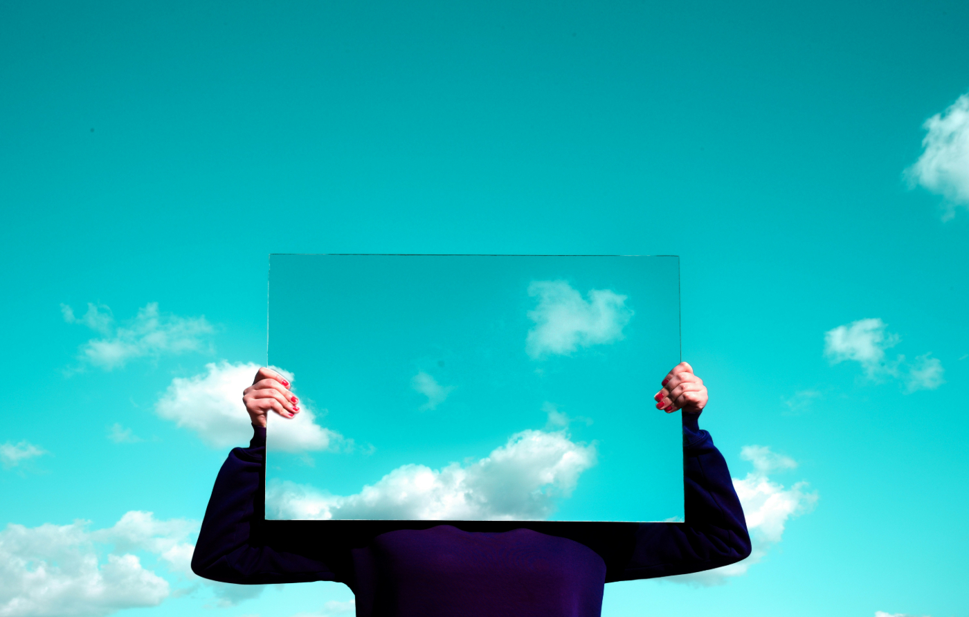 A person holding a mirror In front of their face that is reflecting blue sky and clouds