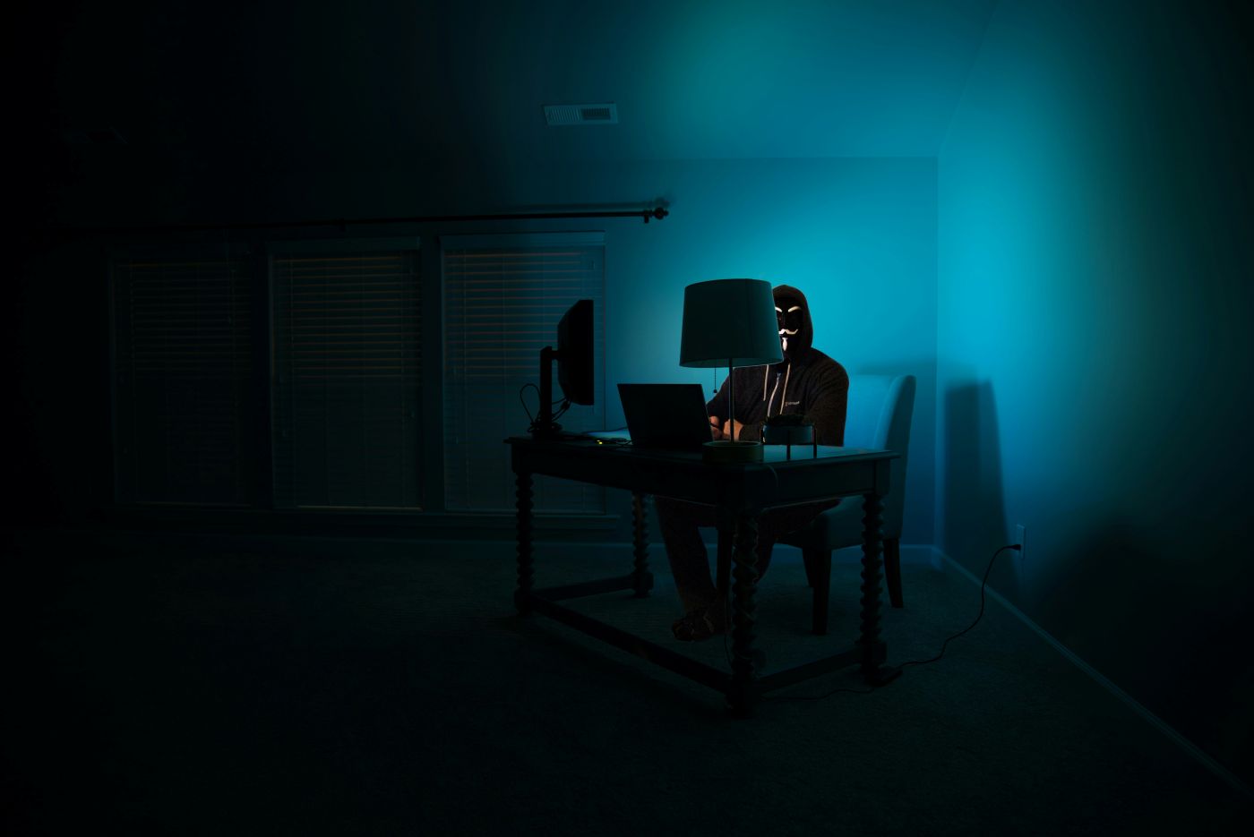 Someone in a mask behind a computer, room lit by blue computer light