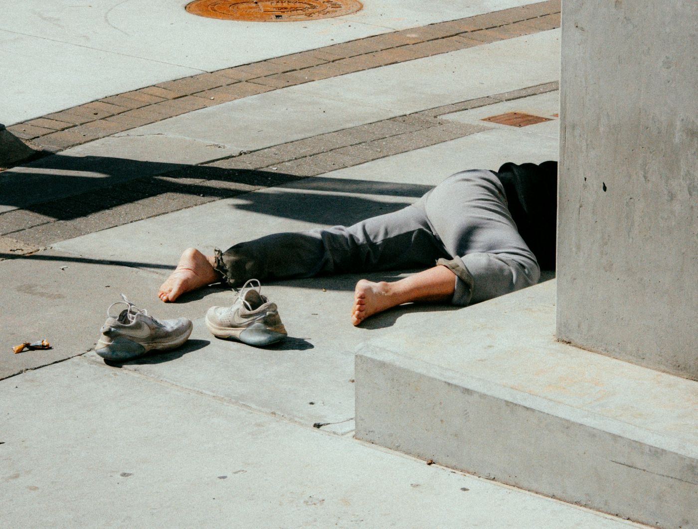A man with shoes off, wearing sweatpants laying on the a sidewalk only half his body visible