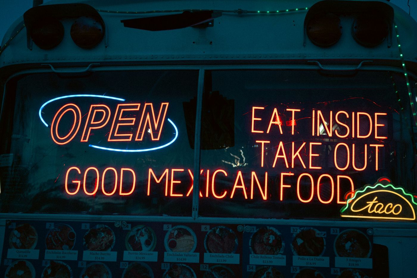 A red neon sign in a window reading Open. Eat Inside. Take Out. GOOD MEXICAN FOOD