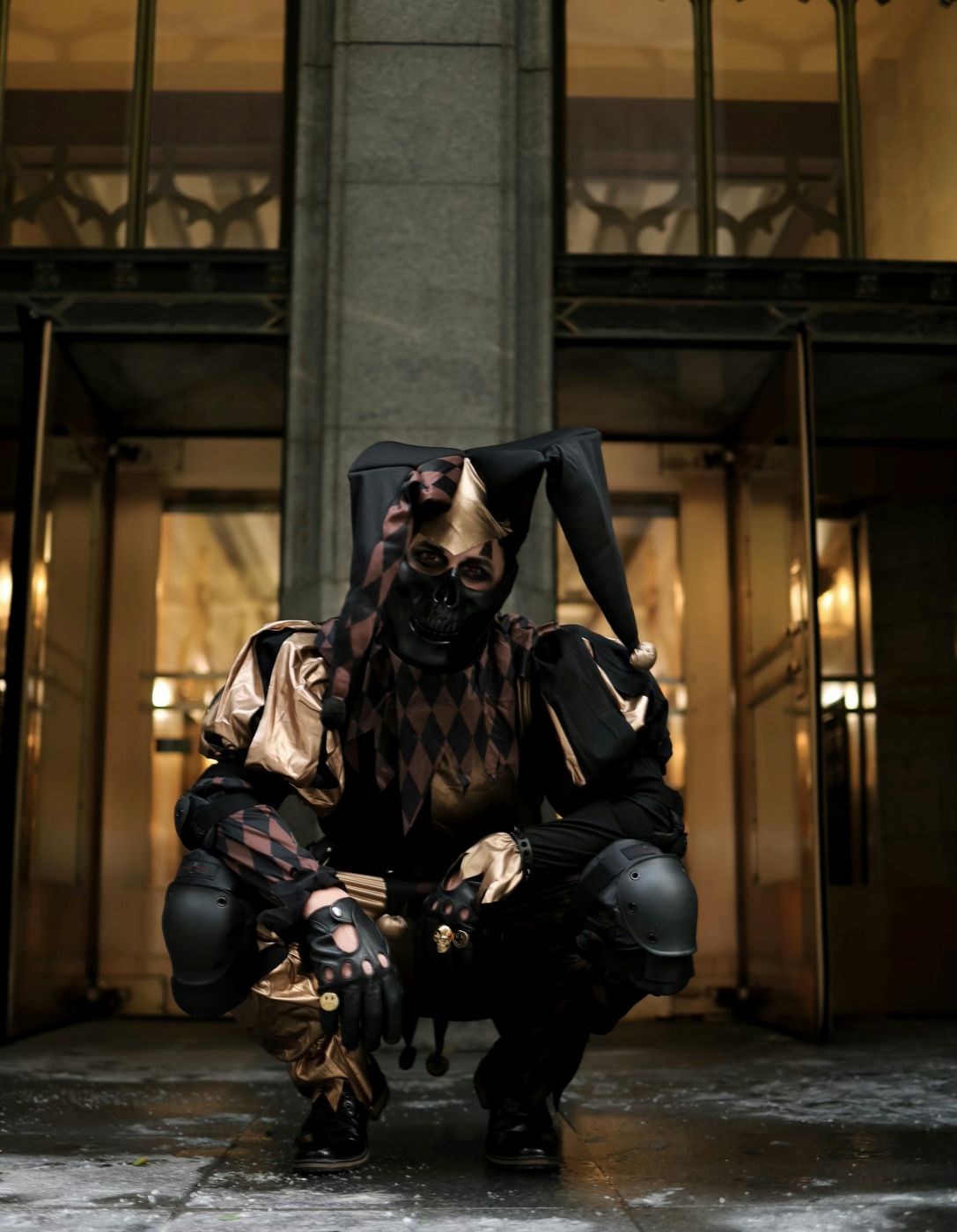 A person in black and gold motley crouching in front of a building