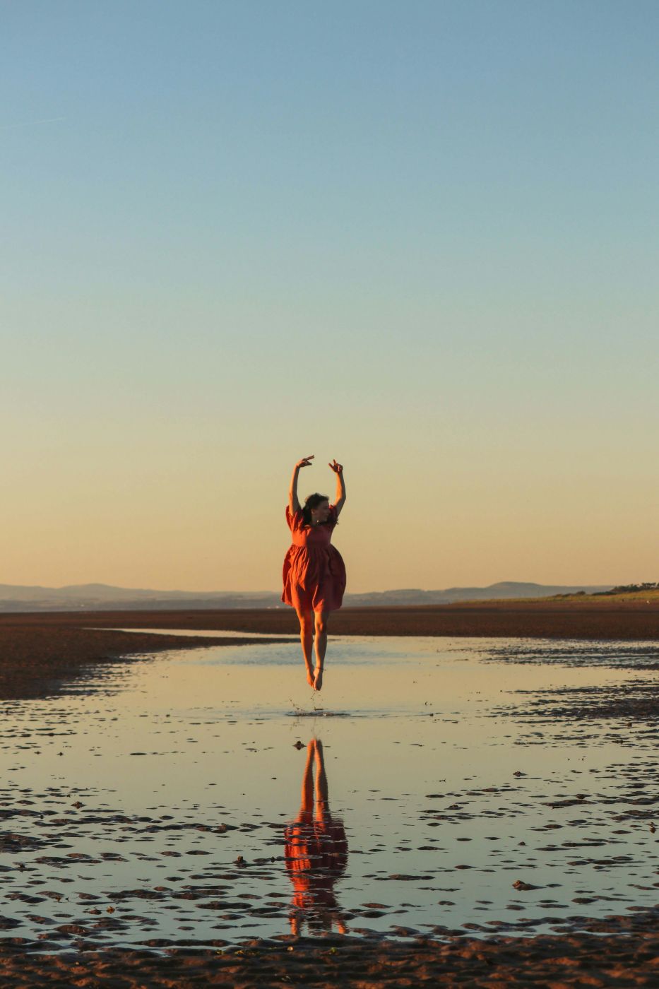A woman in a red dress, mid leap, as she dances on a beach