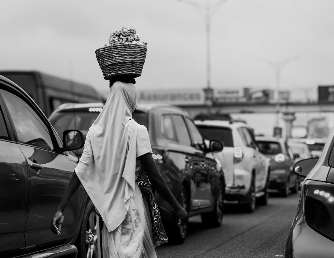 Black and white of a woman with a basket on her head walking beside traffic